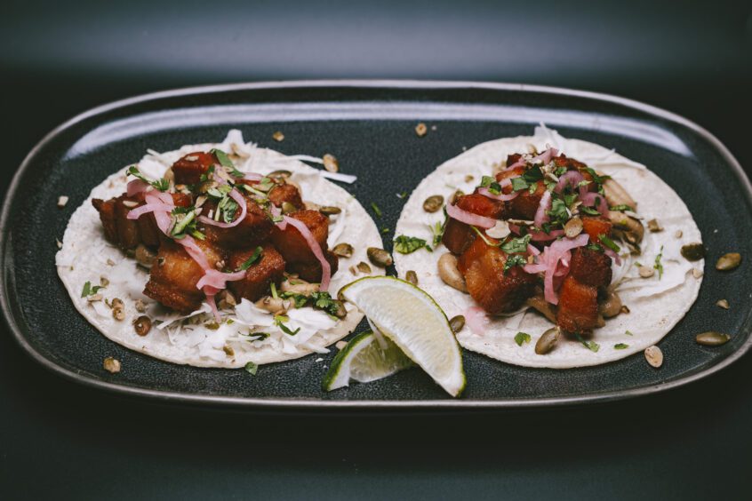 Crispy Pork Belly Tacos on a black plate, topped with cumin-spiced pork, cabbage slaw, salsa macha, pickled onions, pumpkin seeds, and cilantro.