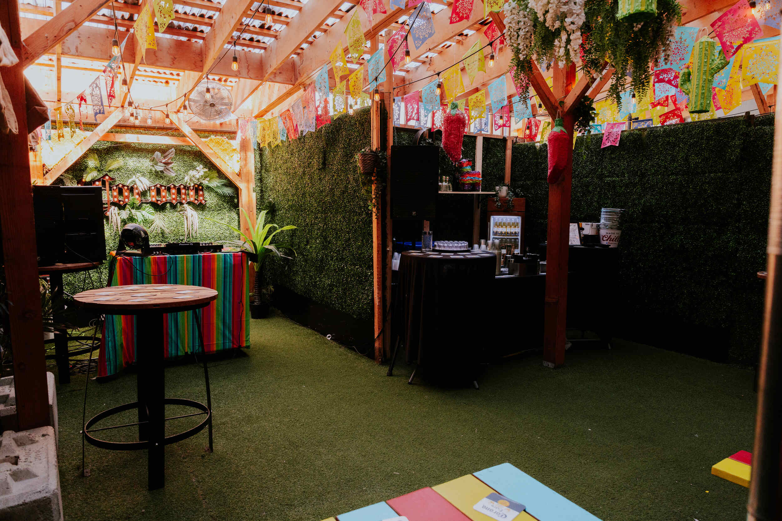 The Pawn Shop YVR's vibrant Hideaway back patio event space. Private bar with DJ booth setup in-front of a Corona sign on a colourful red, yellow, and blue table.