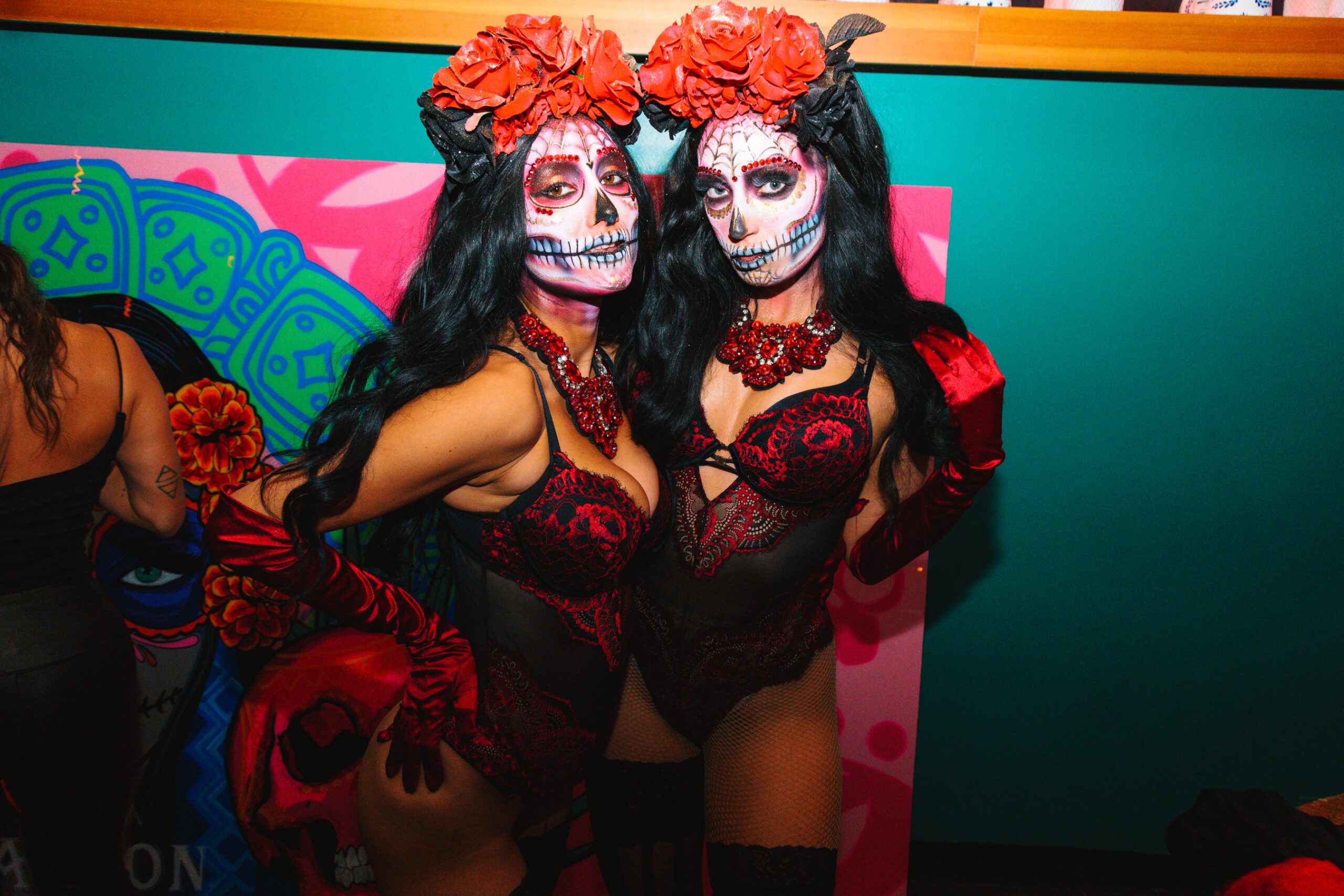 Two dancers in detailed Day of the Dead makeup and red floral headpieces, posing against a colourful mural.
